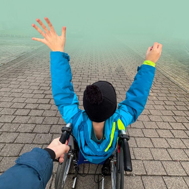 Ein Kind im Rollstuhl hebt auf einem Weg beide Arme.